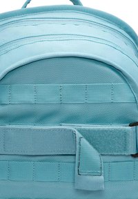 Teal stof rygsæk med en buet top, tekstureret overflade og justerbar rem med velcro. Inkluderer vandret webbing på fronten.