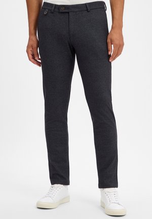 Homme portant un pantalon slim gris foncé et des baskets blanches à lacets, debout devant un fond blanc uni.