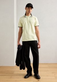 Svetlo žlté polo tričko s čiernymi detailmi, čierne nohavice a čierne topánky. Model drží čiernu mikinu. Čistý, minimalistický dizajn.