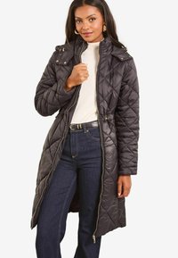 Dlouhý černý prošívaný puffer kabát se zapínáním na zip, kapucí a stahovacími šňůrkami v pase. Má diamantový vzor a zlaté kovové detaily.