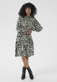 Femeie cu păr creț, îmbrăcată cu o rochie cu mâneci lungi, cu model alb-negru, și cizme negre până la gleznă, stând pe un fundal deschis.