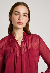 Blusa trasparente bordeaux con pois testurizzati, scollatura a volant, chiusura con bottoni e maniche fluide. Presenta un design a strati e un tessuto morbido.