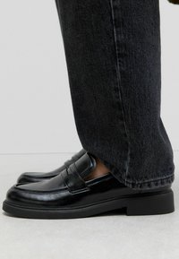 Mocassini in pelle nera con una finitura liscia, dotati di una fascia decorativa lungo il vamp e una suola in gomma nera spessa. Indossati con jeans scuri.