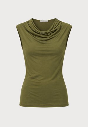 Une femme porte un haut sans manches vert olive au décolleté drapé, un pantalon noir à jambes larges, des chaussures à talons hauts et un grand sac à main noir, sur un fond blanc uni.