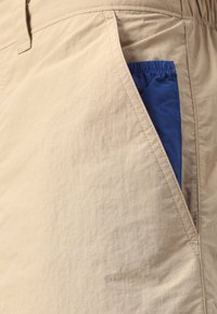 Beige shorts made of lightweight fabric feature a large blue pocket with elastic trim, showcasing a smooth texture and a straight cut design.