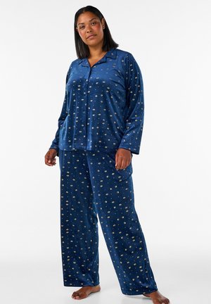 Frau steht barfuß, trägt ein blaues, langärmeliges, geknöpftes Pyjamashirt und passende weite Hosen mit kleinem weißen Punktmuster.
