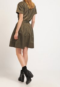 Oliivinvihreä, lyhythihainen mekko, jossa on vyötäröltä kapeutettu malli, liehuva hame ja teksturoidut reunat; yhdistetty mustiin paksupohjaisiin nilkkureihin.
