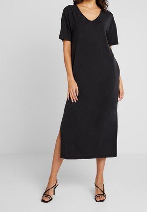 Kobieta ubrana w czarną, midi sukienkę na krótki rękaw z dekoltem w serek i rozcięciami po bokach, w połączeniu z czarnymi sandałkami na wysokim obcasie ze szpagatowymi paskami.