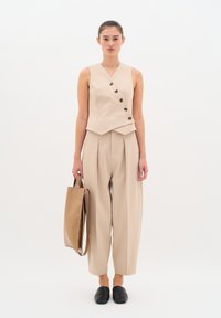 Blusa senza maniche beige con scollatura a V e cinque bottoni neri, abbinata a pantaloni larghi beige e a una borsa a spalla coordinata. Scarpe nere slip-on.