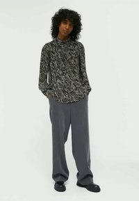 Personne avec des cheveux bouclés portant un haut à manches longues en imprimé zèbre, un pantalon gris ample et des chaussures noires, se tenant avec les mains dans les poches.