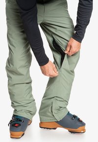 Waterdichte groene skibroek met een ritsventilatie aan de zijkant, gecombineerd met grijze skischolen met een oranje zool en verstelbare gespen.