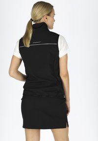 Schwarze ärmellose Jacke mit hohem Kragen, kombiniert mit einem weißen kurzärmligen Hemd. Verfügt über dezentes Branding auf der Rückseite und ein tailliertes Design.
