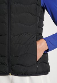 Črn puffer telovnik z oblečenim vzorcem, ki ima sprednji zadrgo in stranske žepe. Oblečen čez modro dolgotrajno majico.