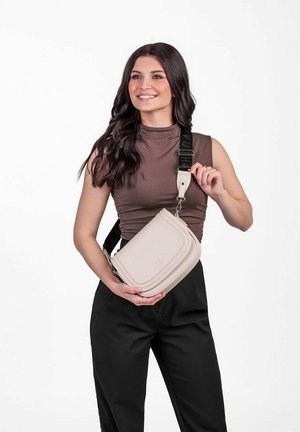 Béžová křížová taška s hladkým povrchem, zakřiveným designem a nastavitelným černým popruhem s bílými akcenty. Modelka drží tašku při pózování.