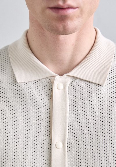 Beige perforált póló, bordás gallérral és három gombbal, textúrált felülettel, kis, háromszög alakú mintákkal.