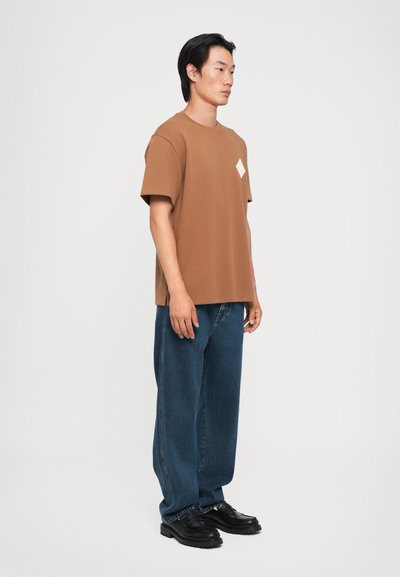 Brown short-sleeve t-shirt with a square patch accent, paired with loose blue denim jeans and black shoes.