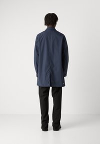 Pomorska vodootporna jakna s ovratnikom, dugim rukavima i ravnim krojem, u kombinaciji s crnim hlačama. Sadrži logo na stražnjem rubu.