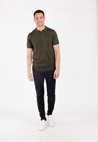 Dunkelgrünes Polo-Shirt mit kurzen Ärmeln, kombiniert mit schwarzen Skinny-Jeans und weißen Sneakern. Das Modell steht vor einem schlichten weißen Hintergrund.