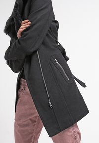 Svart ullblandad kappa med krage i konstpäls, sidoväskor med dragkedja och knytning i midjan. Har rak skärning och en mjuk textur.