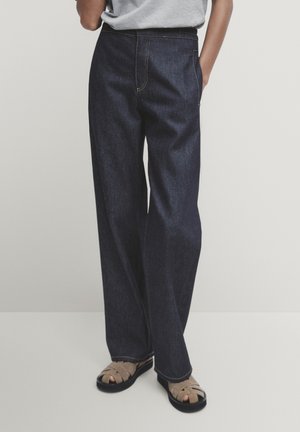 Personne portant un jean en denim bleu foncé ample, un haut gris et des sandales beiges à bouts ouverts, debout avec une main dans la poche.