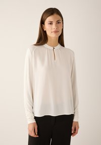 White long-sleeve blouse with a high neckline and a keyhole detail, featuring soft, lightweight fabric and slightly gathered sleeves.