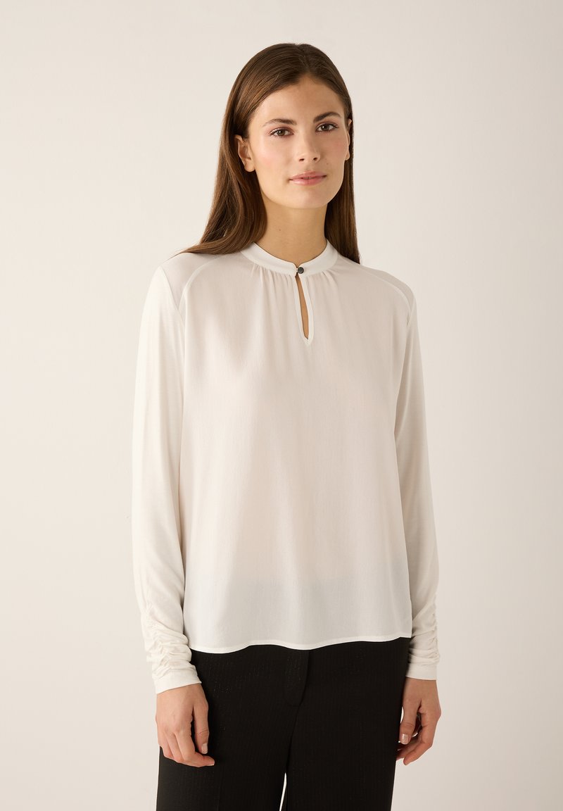 White long-sleeve blouse with a high neckline and a keyhole detail, featuring soft, lightweight fabric and slightly gathered sleeves.