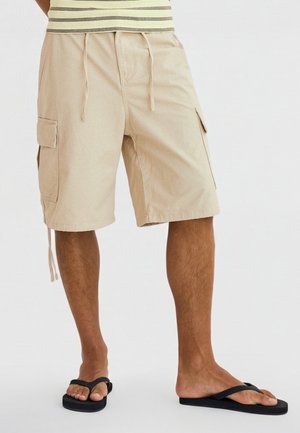 Mann trägt beige Cargo-Shorts mit Kordelzug und schwarze Flip-Flops, steht vor einem schlichten hellen Hintergrund.