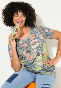 Korte mouw bloemen T-shirt met veelkleurige bloempatronen op een grijze achtergrond; een paar lichtgroene brillen in de mond van het model.