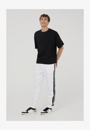 Czarny t-shirt z krótkim rękawem, luźny krój, zestawiony z białymi joggerami z czarnymi paskami po bokach, noszony z czarno-białymi sneakersami.