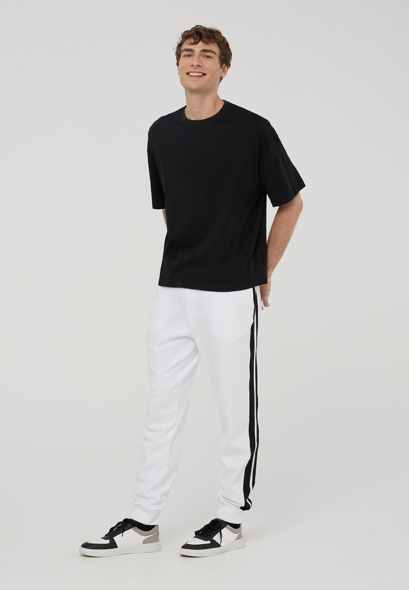 T-shirt nera a maniche corte, vestibilità comoda, abbinata a pantaloni jogger bianchi con strisce nere ai lati, indossata con sneakers nere e bianche.