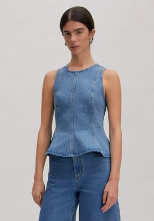 Frau trägt ärmelloses blaues Denim-Peplum-Oberteil und passende hoch taillierte Jeans, steht vor einem schlichten hellen Hintergrund.