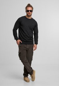 Suéter negro de manga larga con puños acanalados, pantalones cargo de pana en marrón oscuro con bolsillos laterales, y zapatos estilo mocasín en marrón claro.