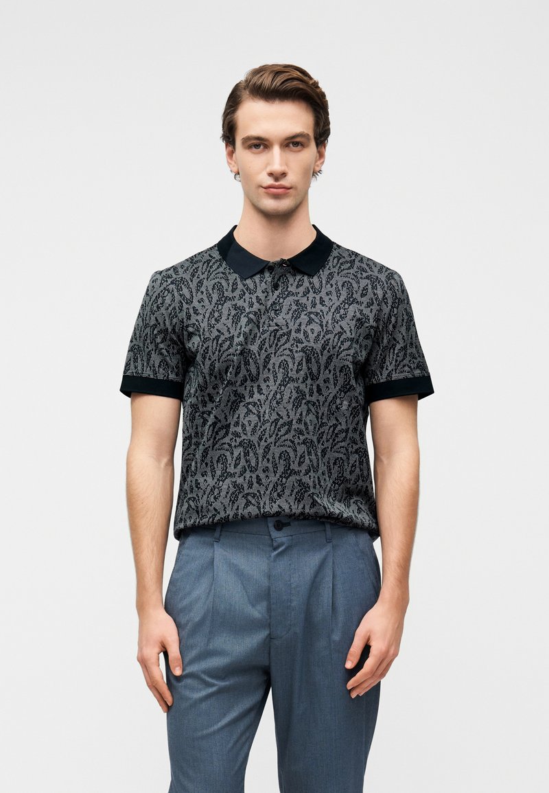 Πολό με μαύρο και γκρι μοτίβο, μαύρο γιακά και κοντά μανίκια, συνδυασμένο με ανοιχτό μπλε παντελόνι. Ομαλή υφή και εφαρμοστό σχέδιο.