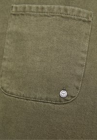 Tissu vert olive avec une poche plaquée à bord arrondi. Petite étiquette logo argentée portant l'inscription « Pacific Republic ». Finition en denim texturé.