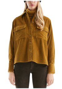 Vrouw die een mosterdgele corduroy overhemd met grote voorzakken draagt, gelaagd over een bijpassende coltrui en donkere broeken.