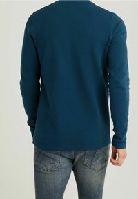 Långärmad, teal ribbad tröja med rund halsringning, tillverkad av textilmaterial. Kombineras med ljusblå denimjeans med bakfickor.