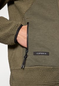 Fleecejacke mit strukturiertem olivgrünem Außenmaterial, ausgestattet mit einer Reißverschlusstasche, schwarzem Besatz und einem seitlich aufgestickten Logo.