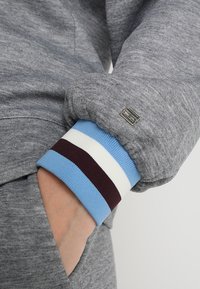Sudadera gris con puños acanalados que presenta rayas en azul, blanco y marrón. Incluye un pequeño logo metálico en el puño.