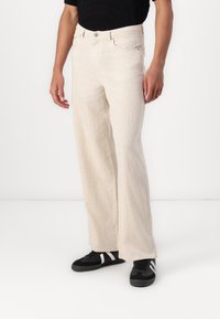 Pantaloni bej cu picior larg dintr-un material texturat, cu talie înaltă, având buzunare frontale și închidere cu nasture, purtați cu adidași negri.