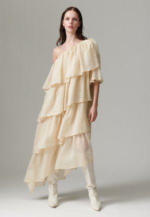 Femme aux longs cheveux bruns portant une robe beige superposée à épaules dénudées et des bottes blanches montantes jusqu'aux genoux, debout devant un fond gris uni.