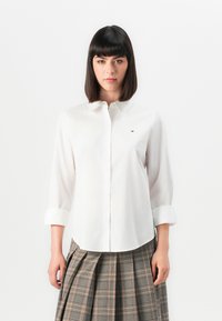 Witte button-up overhemd met een klassieke kraag en oprolbare mouwen, voorzien van een klein geborduurd logo op de linkerkant van de borst, met een soepele textuur.