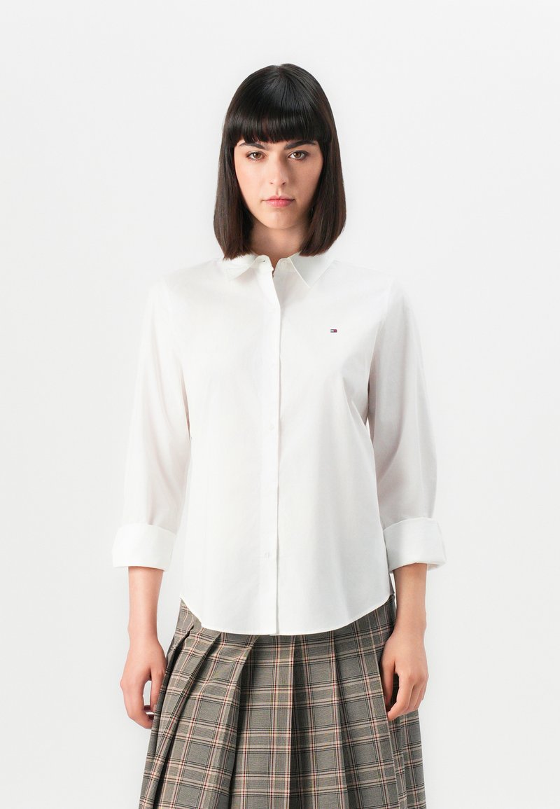 Witte button-up overhemd met een klassieke kraag en oprolbare mouwen, voorzien van een klein geborduurd logo op de linkerkant van de borst, met een soepele textuur.