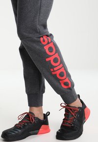 Grijze sweatpants met een groot rood "adidas" logo aan de zijkant, gecombineerd met zwarte sportschoenen met rode accenten en een textuur op het oppervlak.