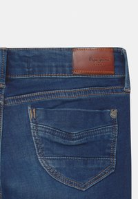 Jeans de mezclilla azul oscuro con una etiqueta de cuero marrón, costuras visibles, un pequeño remache metálico y un bolsillo trasero con diseño curvado.