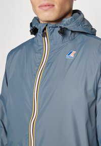 Wasserabweisende, hellblaue Jacke mit Kapuze, Frontreißverschluss und mehrfarbigen Streifendetails. Kway-Logo auf der Brust. Glatte Textur.