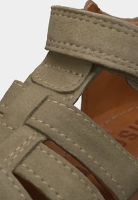 Κοντινό πλάνο με ιμάντες σανδαλιού από μπεζ σουέτ με κλείσιμο βέλκρο και ορατές ραφές, που δείχνει μέρος της καφέ εσωτερικής σόλας.