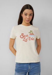 Crème-kleurige katoenen T-shirt met korte mouwen, met de tekst "Sea La Vie" in roze en groen, vergezeld van een palmboom en zonsondergang grafiek.