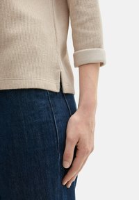 Beige gerippter Strickoberteil mit Dreiviertelärmel, umgeschlagenen Manschetten und seitlichen Schlitzen, kombiniert mit dunkelblauen Jeans.