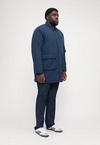 Cappotto imbottito blu navy con chiusura a bottoni, due grandi tasche frontali e maniche lunghe, abbinato a jeans scuri e sneakers bianche.