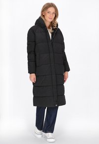 Palton lung negru tip puffer cu glugă, având un design matlasat, buzunare laterale și închidere cu nasture la tiv. Purtați peste blugi albaștri.